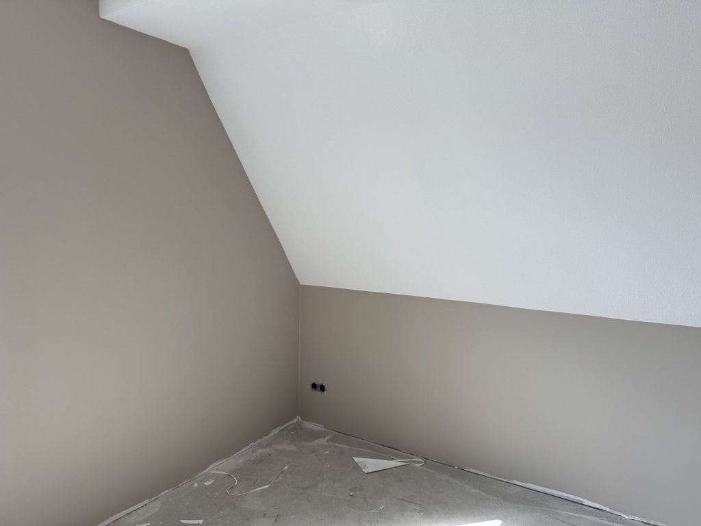 Zolderkamer in een nieuwbouwwoning met een schuine wand, lichte wanden en een betonvloer vol snippers
