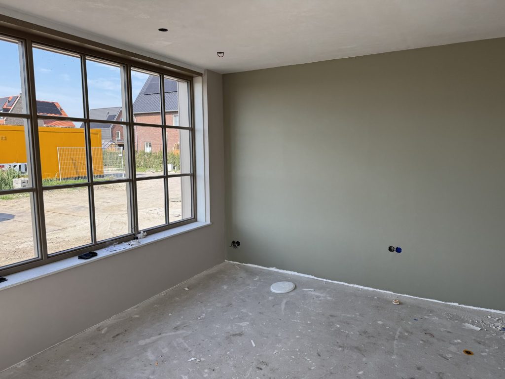 Nieuwbouwkamer met een egaal afgewerkte grijsgroene wand en een groot raam met roedes naar een bouwterrein