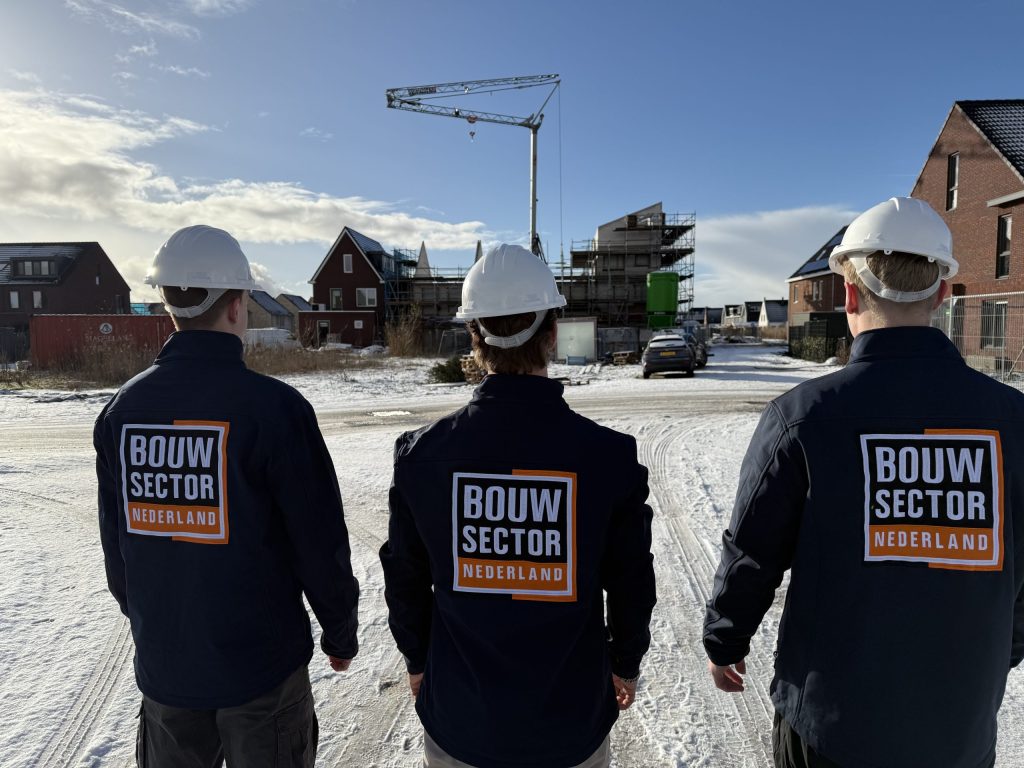 Drie medewerkers met witte helm en jas van Bouwsector Nederland lopen over een besneeuwde bouwplaats naar nieuwe huizen en een bouwkraan